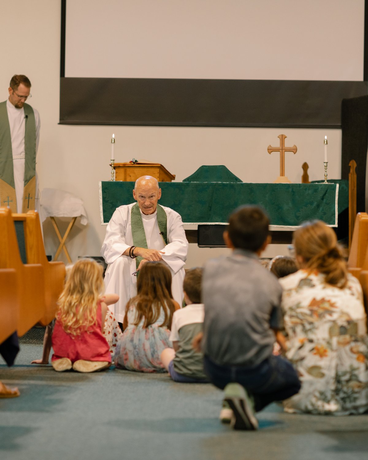 The rector seated, giving the children's homily as kids gather on the floor.