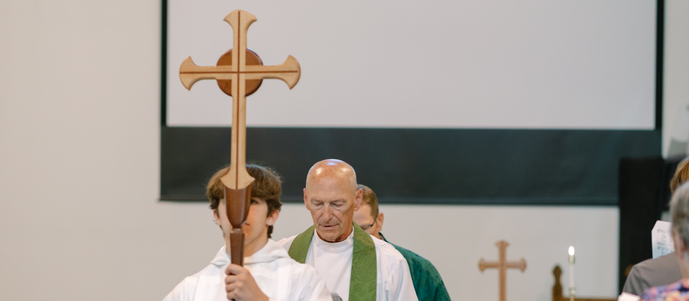 An acolyte carries the processional cross down the aisle, followed by the rector in a green stole.